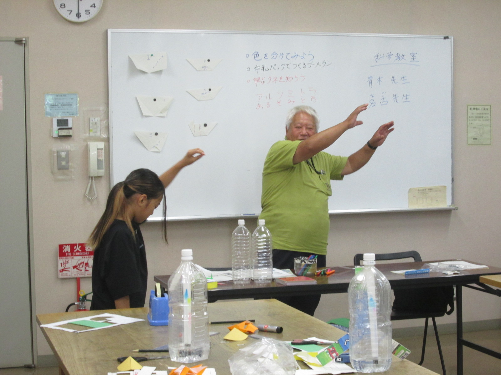 男性と少女が室内で両腕を広げており、ホワイトボードには日本語で「科学教室」や先生の名前が書かれている写真