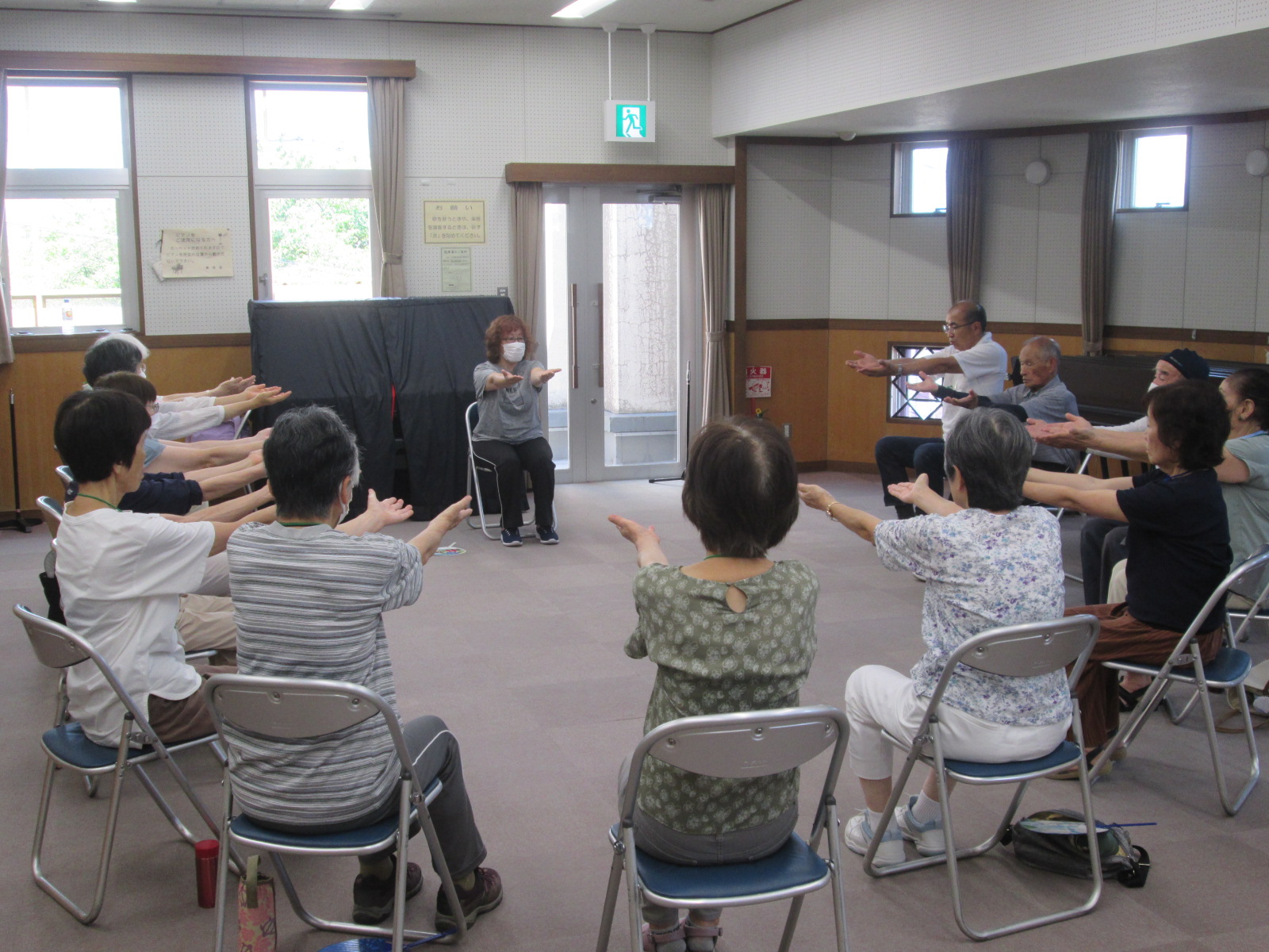輪になって椅子に座った高齢者が、講師の指示に従い、両腕を前に伸ばす体操をしている様子を捉えた写真