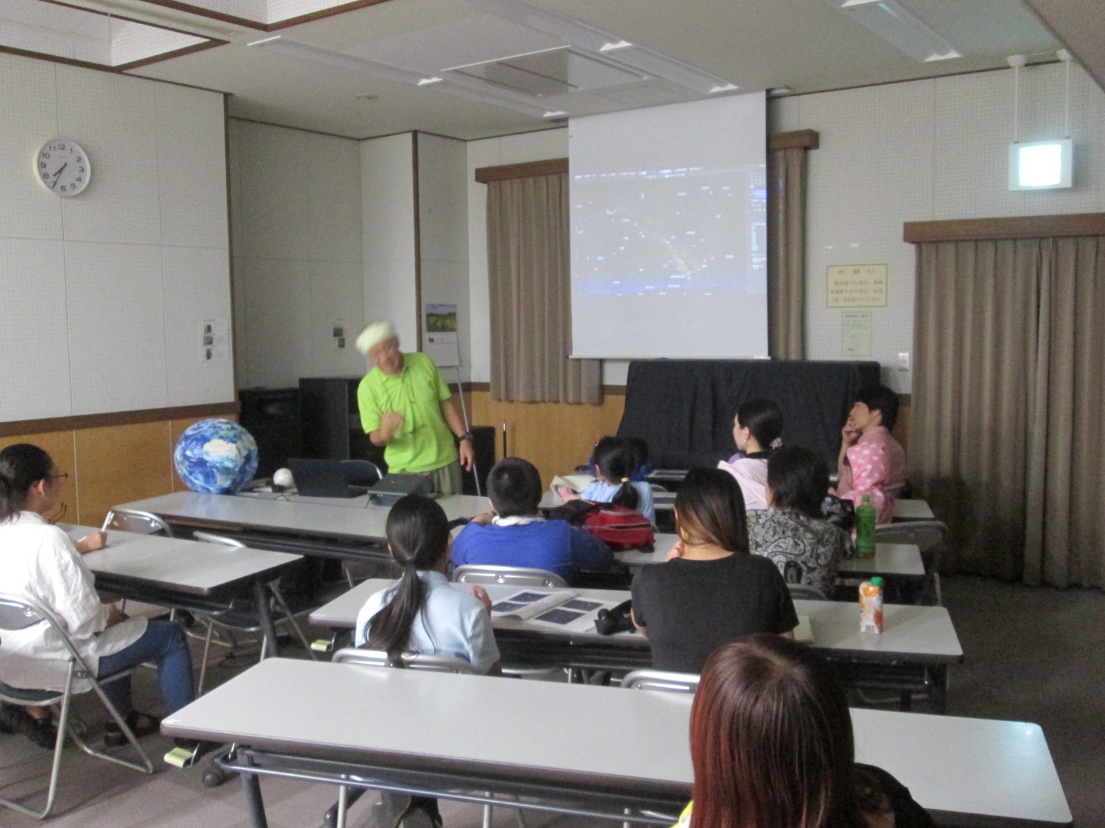 講師が蛍光グリーンのTシャツを着て教卓の前に立ち、スクリーンに映し出された星の映像を使い、机に座った子供たちと保護者に対し熱心に天体観測について解説している様子の写真