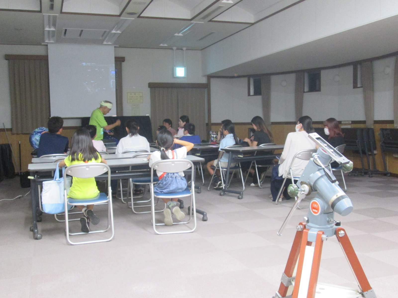 部屋の前方に立つ講師が子どもや保護者に向けて話しており、手前には天体望遠鏡が設置された学習会の風景を写した写真