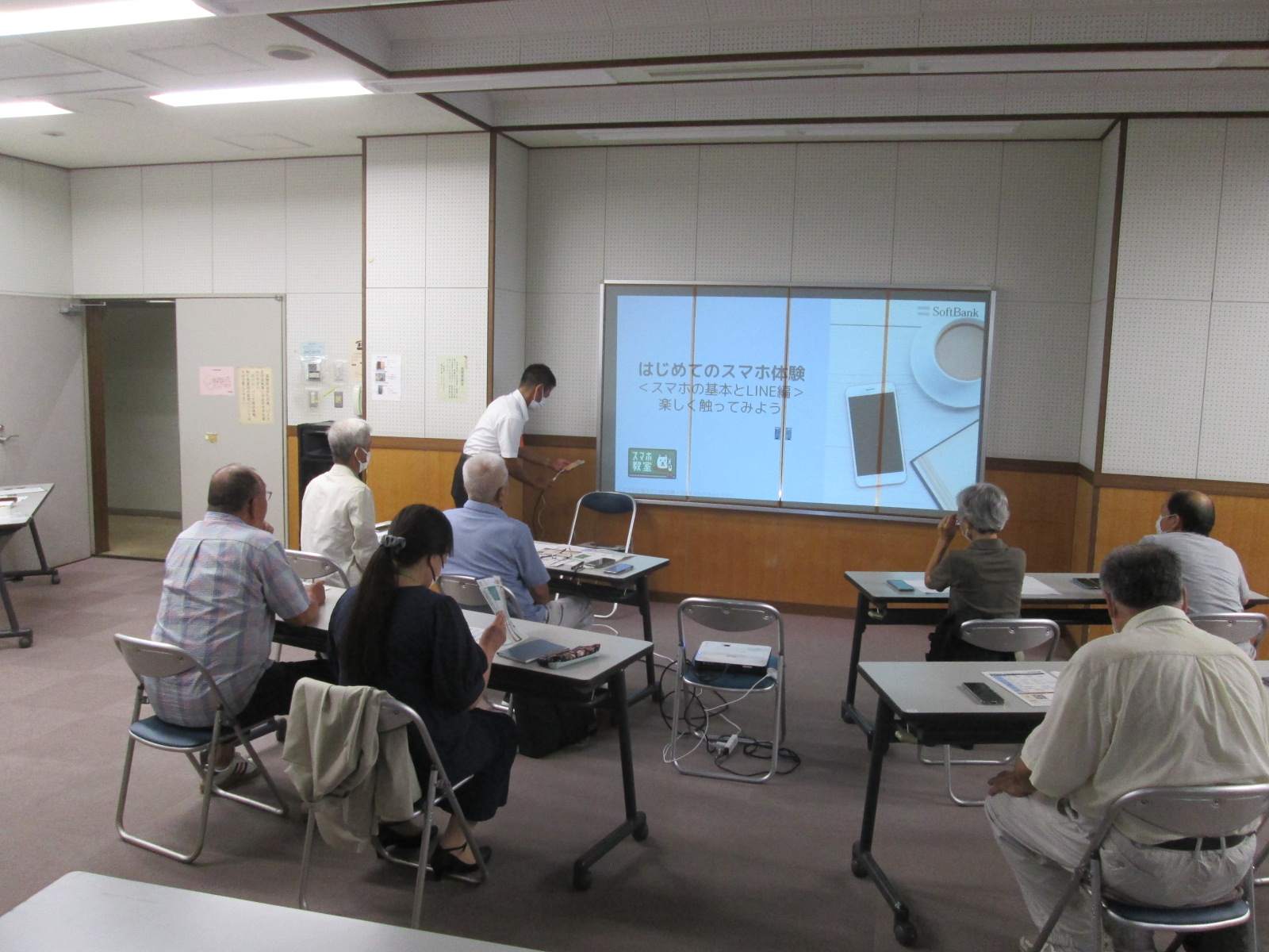 高齢者たちが会議室でテーブルと椅子に座り、プロジェクターに映し出された「はじめてのスマホ体験」という画面を見ながら、スマートフォン教室に参加している様子の写真