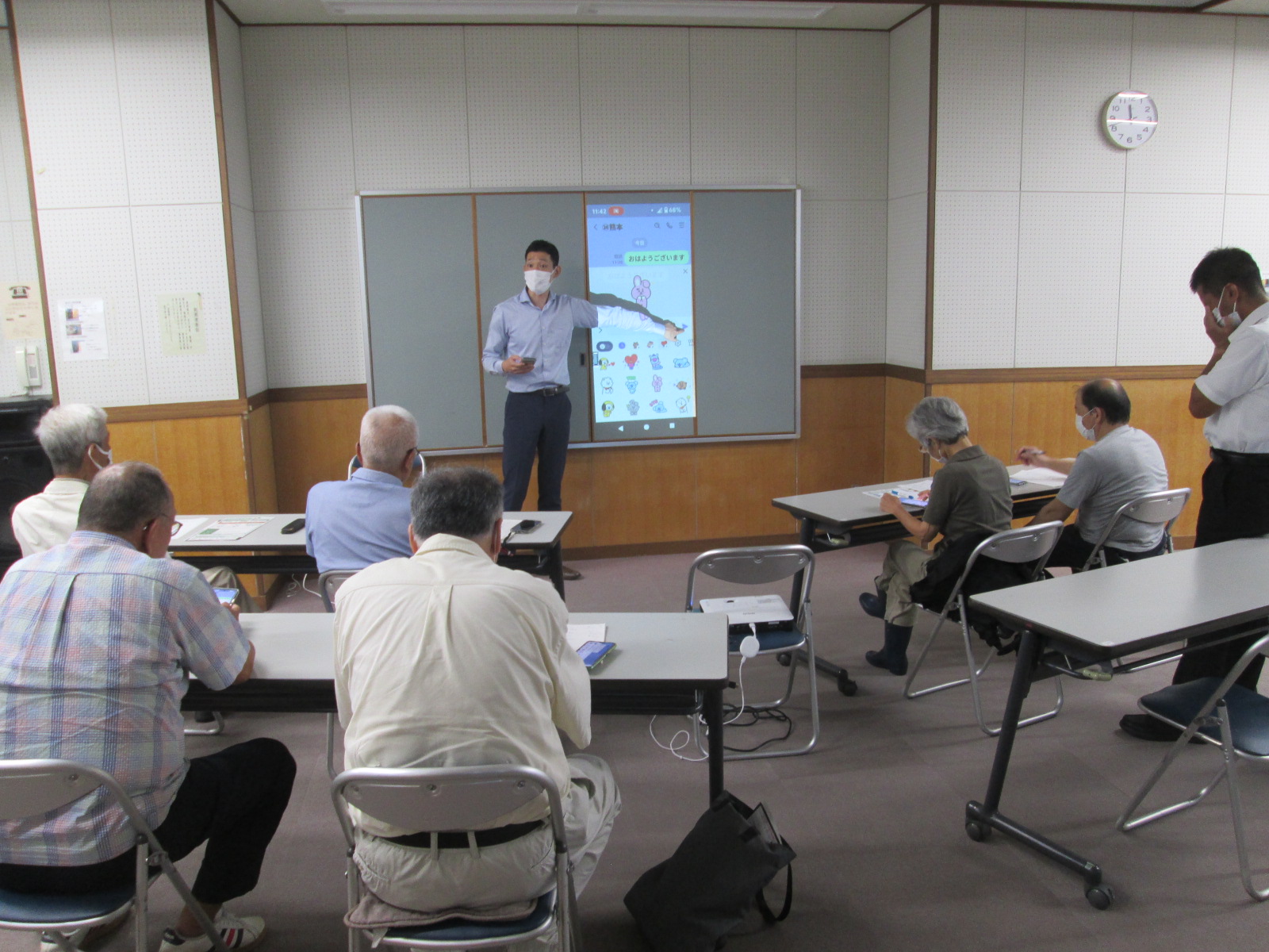 椅子に座った高齢者たちが前方のプロジェクターに映し出されたスマートフォンの画面を集中して見ており、講師がマイクを持ちながら指差し説明を行なっている写真