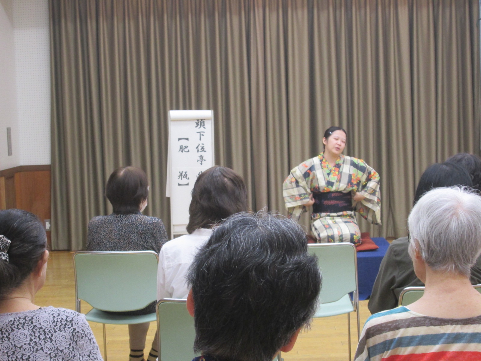 和服を着た女性が舞台に正座し、複数の参加者に向かって落語を披露している様子を撮影した写真