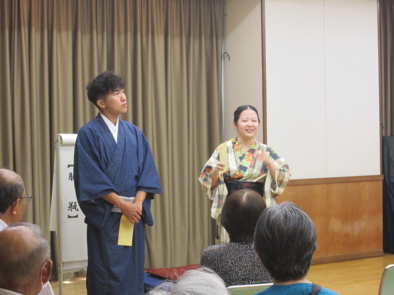 ステージ上で和服を着た男女が、落語を披露している様子を、観客席側から撮影した写真