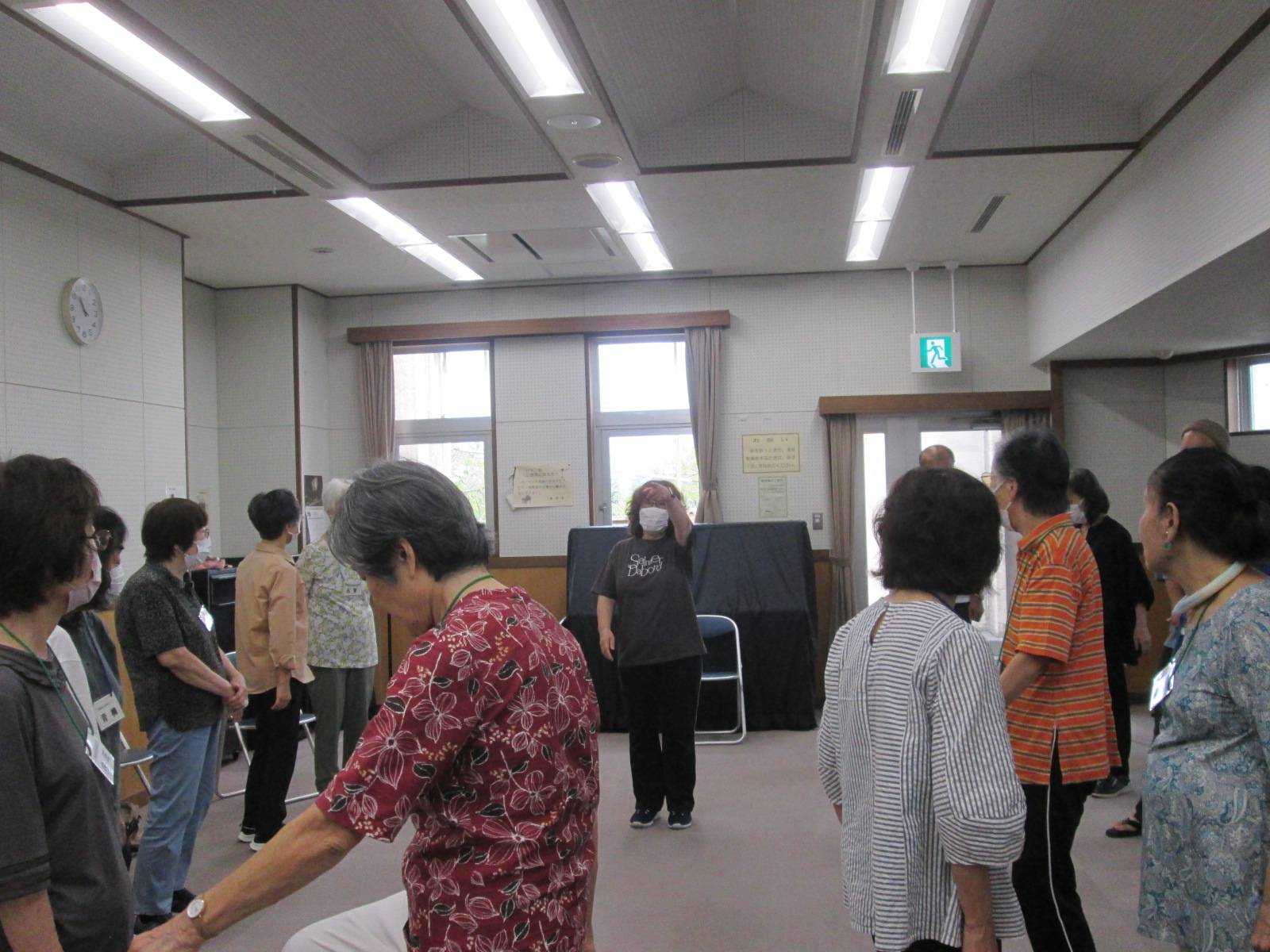 白い壁の広間で、複数人が集まり、マスクを着用した講師の女性の指導を受けている体操の様子の写真