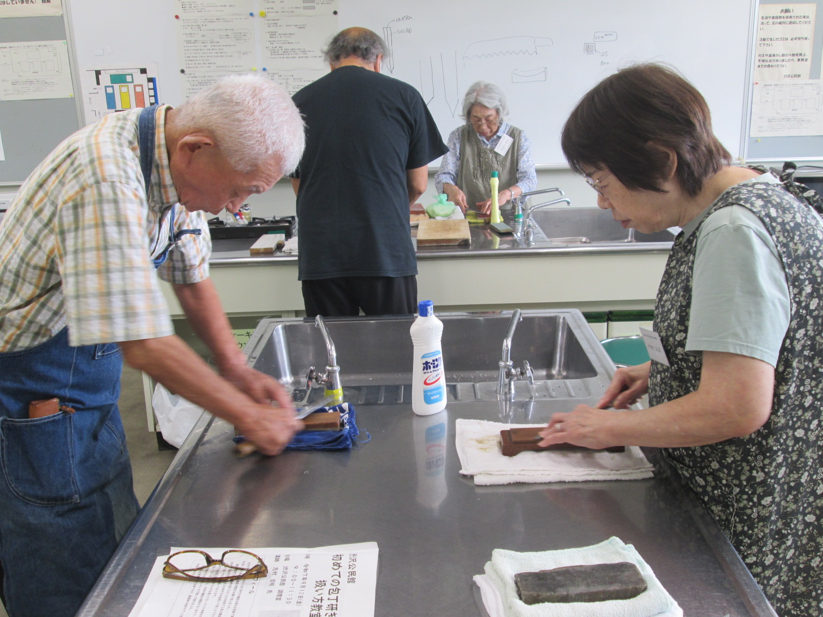 調理実習室のステンレス製の作業台で、エプロンを着用した男女2人が、それぞれ濡らした砥石を使って包丁研ぎの実習を行っている様子の写真