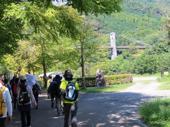 太陽光が降り注ぐ中、リュックサックを背負った数人のハイキング参加者が、木々に囲まれた舗装路を歩いており、その先に大きな吊り橋が見える景色の写真