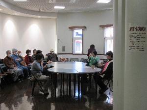 数人の参加者が円形のテーブルを囲んで大きな茶碗からお茶を飲んでおり、後方で複数の参加者が椅子に座り様子を見守っている写真