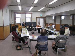 机を口の字に並べた席に座った参加者たちに、一人の女性が資料を配る様子を捉えた写真