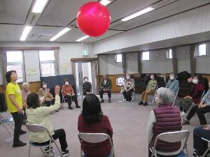 円形に並べた椅子に座った参加者たちが、大きな赤いボールをバレーのように上にあげる遊びをしている様子の写真