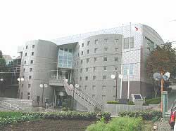 曲線的なデザインが特徴的なコンクリート造りの建物で、前面に大きな階段があり、多数の小さな正方形の窓が等間隔に配置された渋沢公民館外観写真