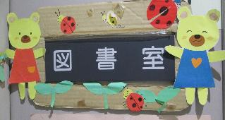 ボール紙の台紙に黒い紙が貼られ、白抜きで「図書室」と書かれたプレートの周りを、赤いワンピースと青いサロペットを着た二匹の黄緑色のクマの切り紙や、テントウムシと葉っぱのモチーフで装飾した、愛らしい掲示物の写真