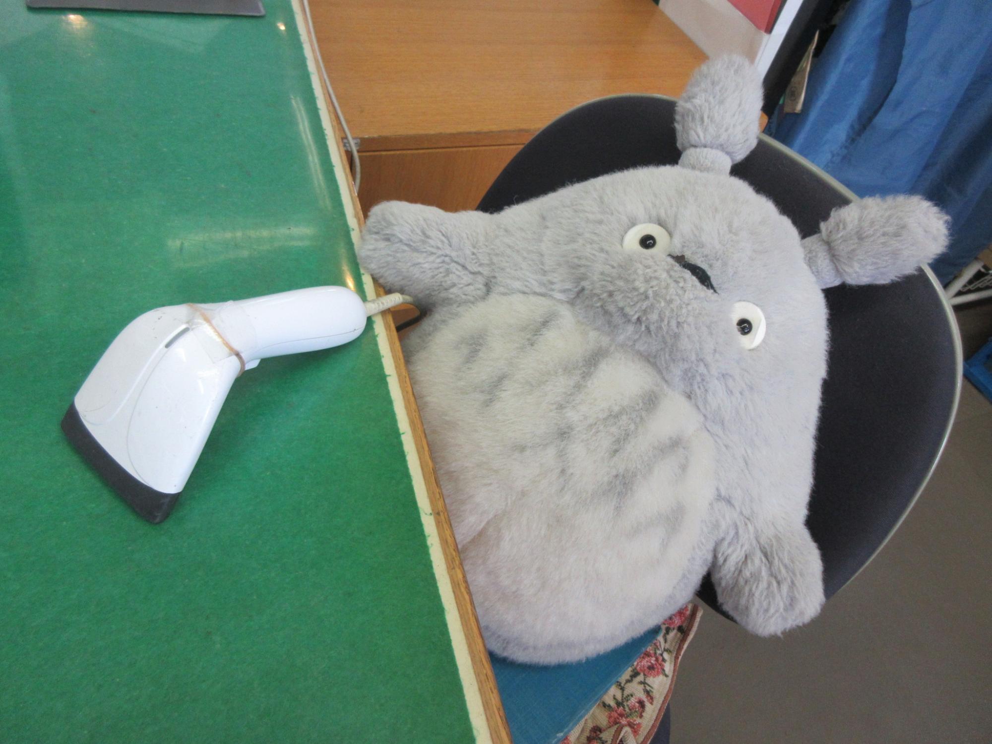 緑色のカウンターの上に白いバーコードリーダーが置かれ、その隣の椅子にはグレーの毛並みと大きな目を持つぬいぐるみが座っている様子を上から捉えた写真