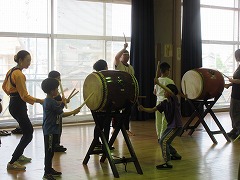 広い室内で、指導者と数名の男の子たちが、バチを両手に持ち2つの大きな和太鼓をそれぞれバチで叩いている写真