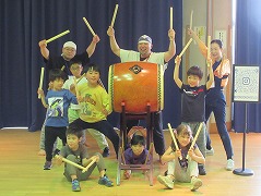 和太鼓を中心にして、指導者と思われる大人3名と、複数の子供たちが、バチを高く掲げたり構えたりしながら笑顔でポーズをとっている集合写真