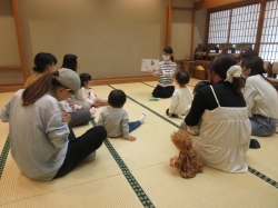 畳敷きの和室で、中央の女性が絵本を広げ、円になって座る大人数名と幼児数名に読み聞かせをしている様子の写真