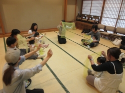 畳敷きの和室で、中央の講師らしき女性を囲み、黄緑色のシフォンスカーフを両手で持ってあげて親子が遊んでいる様子の写真