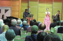 ステージ上でピンクの華やかなドレスを着た女性が歌唱しており、後方には男性のコントラバス奏者と女性のバイオリン奏者、左端にはピアニストがおり、参加者が熱心に鑑賞している様子の写真