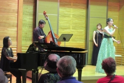 コンサート会場のステージ上で、緑色のロングドレスを着た女性歌手が歌唱しており、コントラバス、ピアノ、バイオリンの奏者たちが演奏する様子を、参加者の後方から写した写真