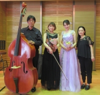演奏会後のステージで、左からコントラバス奏者の男性、バイオリン奏者の女性、薄紫のドレスを着て花束を持った歌手の女性、ピアニストの女性の計4名が立って並んでいる集合写真