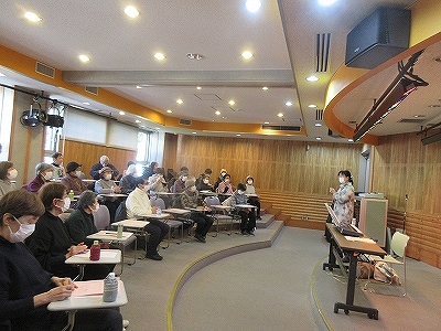 講師らしき女性が壇上で参加者を前に話しており、参加者は講義室の机に座ってマスクを着用し、資料に書き込みながら真剣に聞いている様子の写真