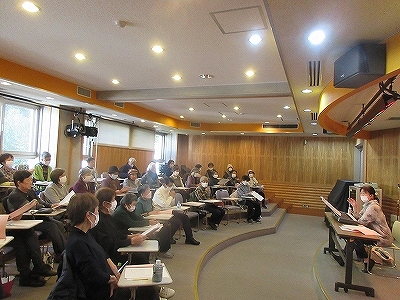 講義室の半円状に配置されたデスクに多数の参加者が座り、全員マスクを着用して手元の資料に目を通しており、講師が電子ピアノが置かれた机に座り説明をしている写真