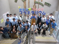 「はだのみんなでプラネタリウム」と書かれた壁画の前で、20名以上の参加者が、階段に集合して笑顔でカメラに向かってポーズをとっている様子の写真