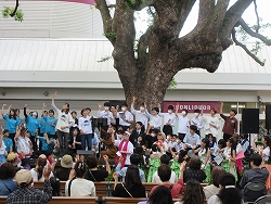 屋外のイベント会場で、大きな木の根元にあるステージと観客席に分かれ、ステージ上の大人と子ども、観客の多くが両手を挙げて歓声を上げているような様子の写真