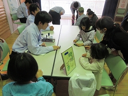 会場の長テーブルを囲んで、学生らしき数人の大人と、大人に教えられながら作業をする小さな子どもたちが、卓上の教材を使って学習や交流をしている様子の写真