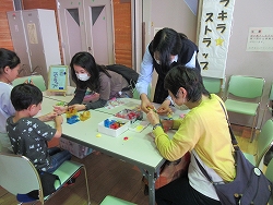 会場の机を囲んで、数人の子どもと大人が座り、カラフルなビーズのような小さなブロックを使い、スタッフらしき人が指導しながら、手作り体験している様子の写真