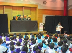 広い部屋で、黒い幕の前にいる演者たちが動物などの被り物をして人形劇のようなパフォーマンスを行い、多くの園児たちが床に座ってそれを見ている写真