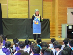 ステージ上で、エプロンを着用した年配の女性が、黒い幕の前に立ち、床に座っている大勢の子どもたちに向かって、何かを手に持ちながら楽しそうに話しかけている写真