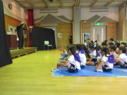 体育館のような会場で、床に座って行事に参加している多数の子どもたちに、ステージ上で黒い衣装を着た方が何かの道具を持って見せている写真