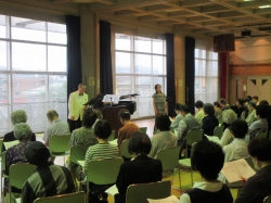 大きな窓のある広い室内で、グランドピアノと譜面台を前に、二人の指導者が多数の参加者に合唱のl講習をしているのを後ろから写した写真