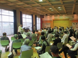 大きな窓のある広間で、多数の参加者が薄緑色の椅子に座り、前方でグランドピアノと二人の指導者による合唱または音楽の指導を受けている様子を後方から捉えた写真