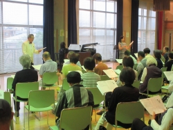 広々とした明るい室内で、大勢の参加者が薄緑色の椅子に座り、それぞれ楽譜を手に持って、グランドピアノを挟んで立つ男女二人の指導者から音楽の指導を受けている様子の写真