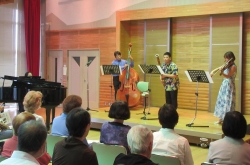 ステージ上で、グランドピアノの近くで男性がコントラバス、男性が管楽器、女性がヴァイオリンを演奏する三人組の楽団が、客席の聴衆を前にコンサートを行っている様子の写真