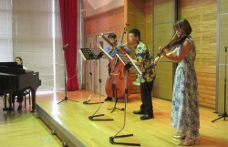 明るいステージの上で、女性がヴァイオリン、男性がコントラバス、もう一人の男性が木管楽器を演奏しており、左奥にはグランドピアノが配置されている室内楽コンサートの様子の写真