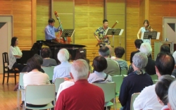 ステージ上で、グランドピアノの周りにコントラバス、木管楽器、ヴァイオリンを演奏する三人の楽団と聴衆が座り、室内楽コンサートの鑑賞風景を捉えた写真