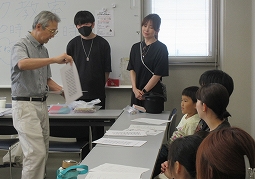 教室のような場所で、一人の年配の男性講師が数枚の資料を配りながら、何人かの若者や子ども連れの家族に向けて説明を行っている様子の写真