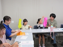 テーブルを囲んで座る大人数名と子どもが、それぞれ黄色やピンクなどの鮮やかな色の布を持ちながら、手品の練習をしている様子の写真