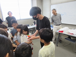 男性の指導者が大勢の子どもたちに囲まれながら、手に持った小さなブロックを真剣な表情で見つめる女の子に見せながら手品をしている様子の写真