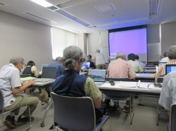 パソコンが並ぶ教室で、主にシニア世代の参加者がそれぞれのノートパソコンに向かい、講師と思われる人物がスクリーン近くに立って指導している写真