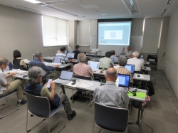 会議室のような部屋で、主に中高年以上の参加者たちが各自のノートパソコンを操作し、スクリーンに映し出された画面を見ながら一人の講師から講義を受けている写真