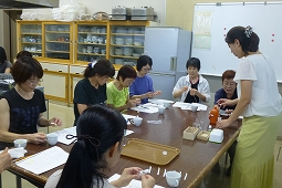 机を囲んで複数の女性たちが座り、講師がアロマオイルを作っている過程を説明しながら参加者と一緒に作業をしている写真