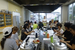 広い料理場で参加者たちが長いテーブルを囲んで座り、前に立っている講師がアロマオイルの作り方の説明をしている様子の写真