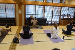 前に座っている先生を見ながら、畳の部屋で複数の人がヨガマットを敷いて座り、ヨガをしている様子の写真