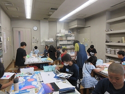 室内で、複数の子どもたちが各自の机に向かって絵を描いたり工作をしたりしており、講師が指導や手助けをしている様子の写真