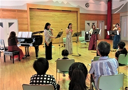 ドレスを着た女性たちがピアノやアコーディオン、フルートなどの楽器を演奏し、観客が座って聴いているところを横斜めから撮影した写真