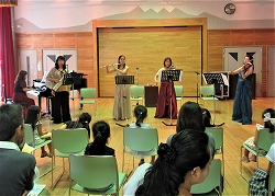 ドレスを着た女性たちがピアノやアコーディオン、フルートなどの楽器を演奏し、観客が座って聴いているところを正面めから撮影した写真